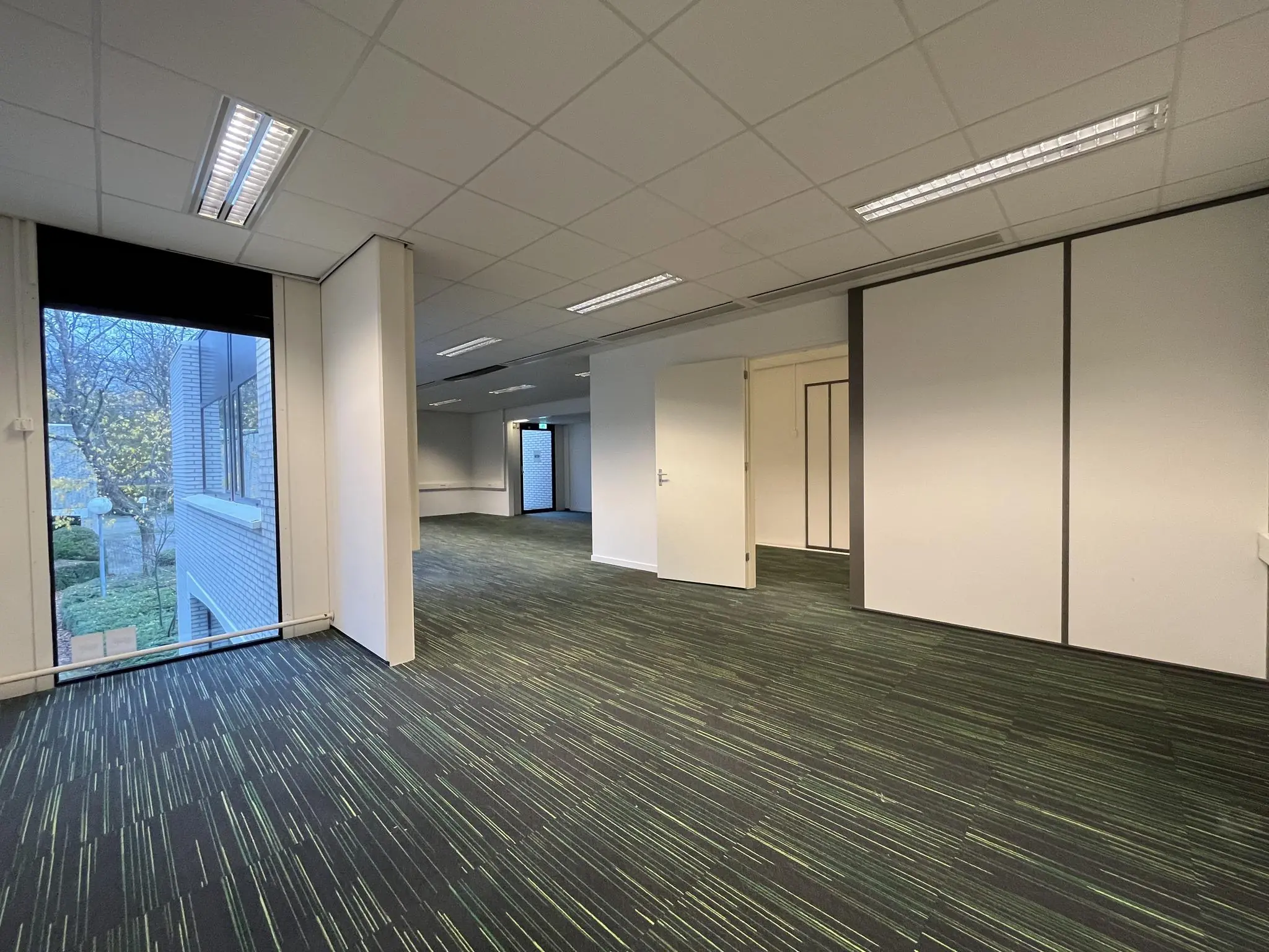 Leegstaand kantoorruimte in het pand aan de Burgemeester Verderlaan met moderne verlichting en groen gestreept tapijt.